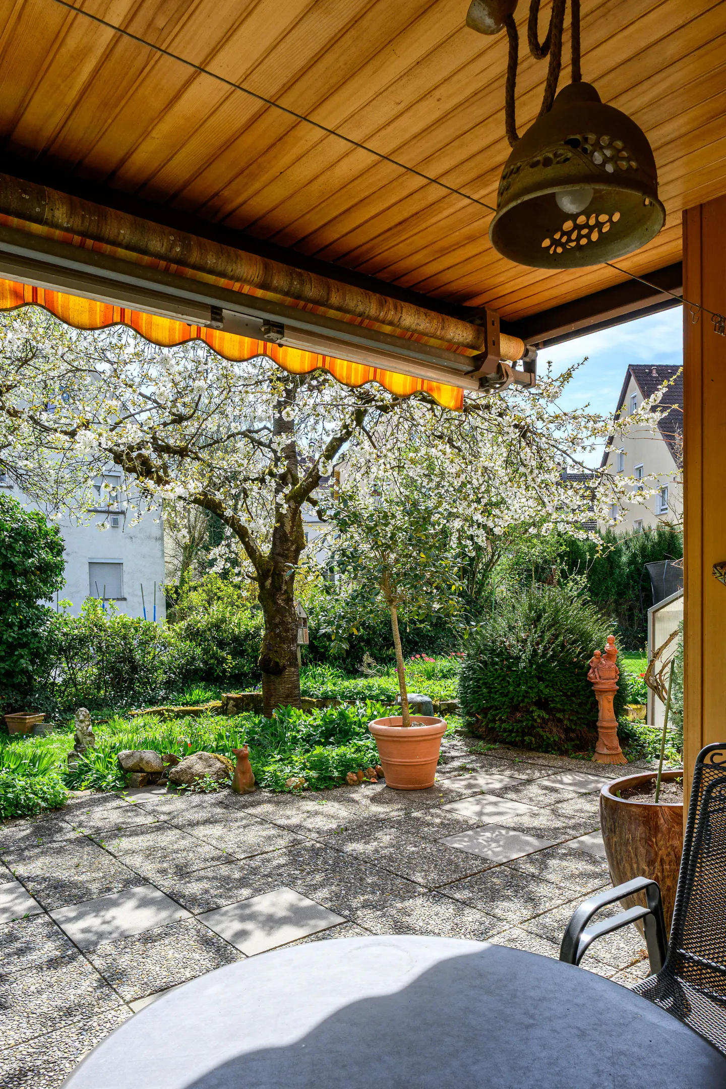 Covered patio with a view of a garden with flowering trees and green bushes. A light fixture hangs from the wooden ceiling.