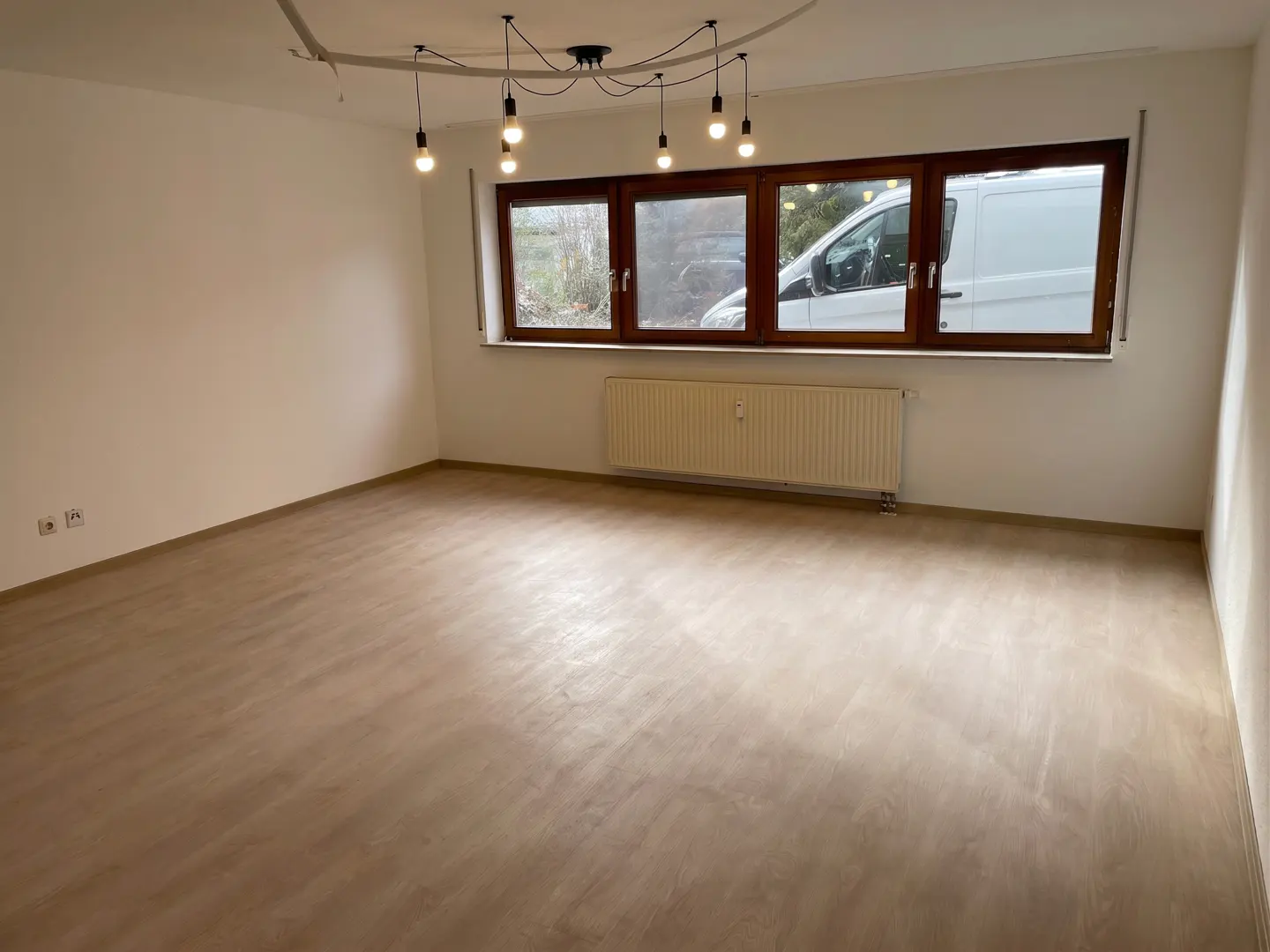 Bright, empty room with light wood floors, white walls, and a modern light fixture. A window shows a white van outside.