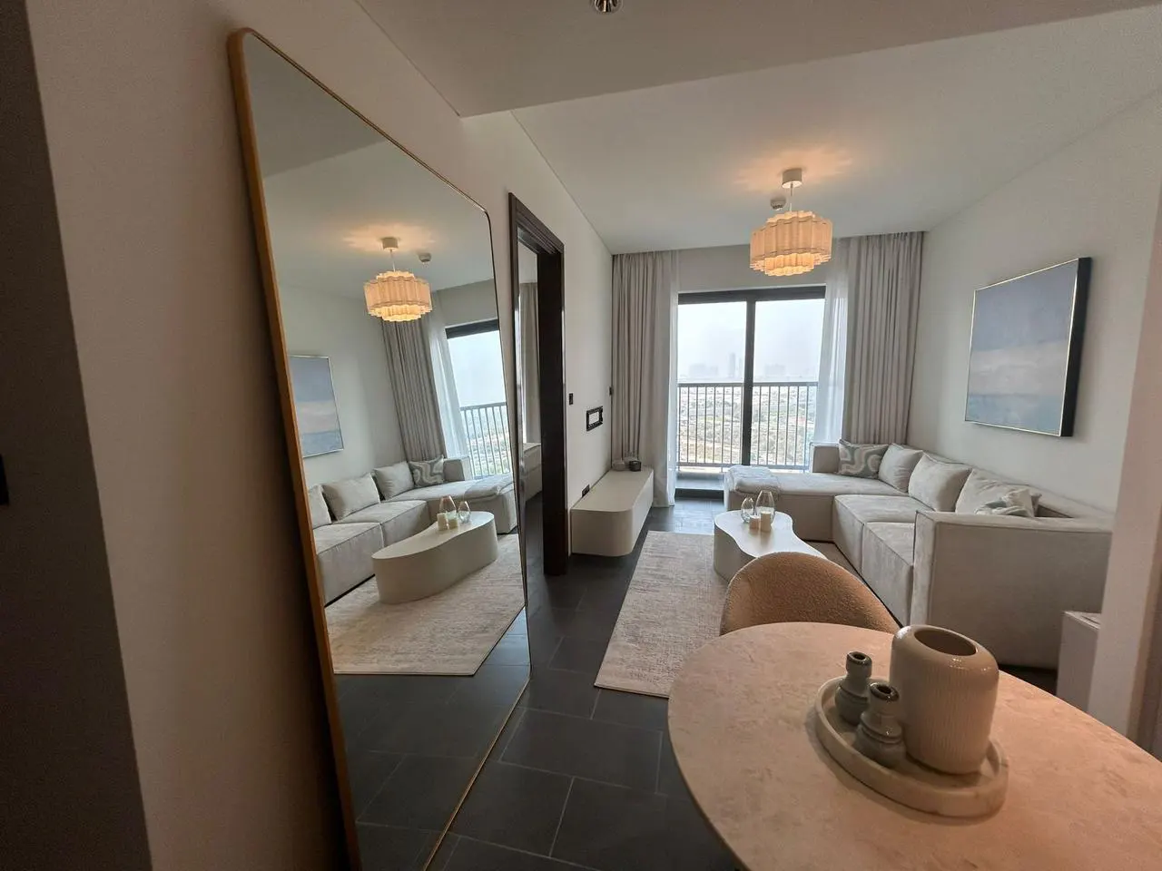 Living room with gray sectional sofa, white rug, and coffee table. A large mirror reflects the room. Balcony with city view.