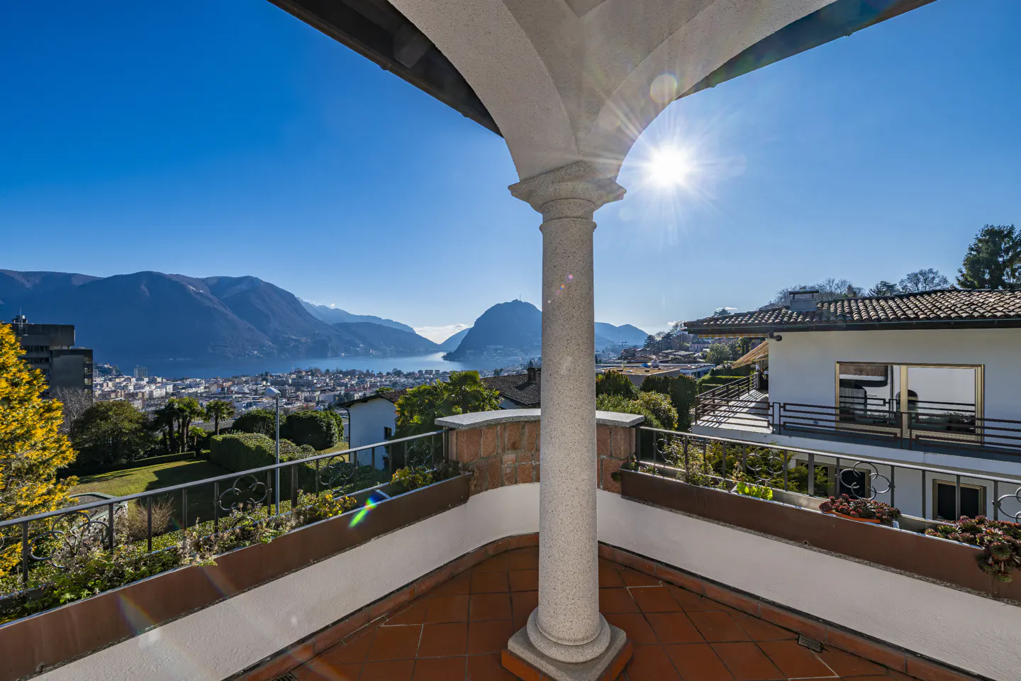 View from a terracotta-tiled balcony with a stone column, overlooking a city, lake, and mountains under a bright blue sky.