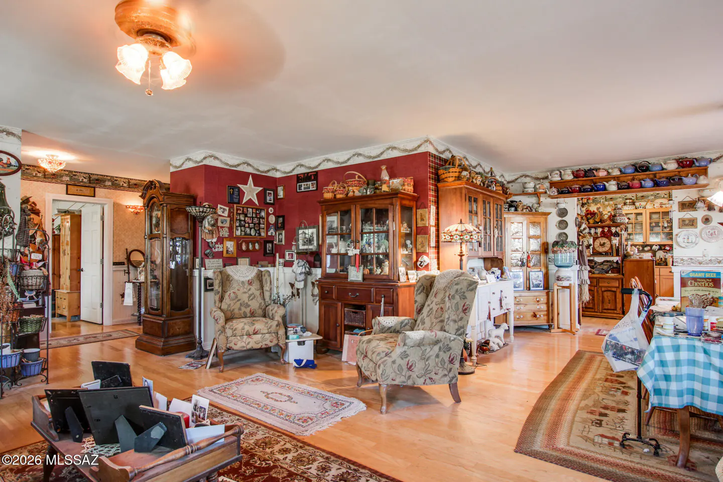 A cozy living room with hardwood floors, floral armchairs, and antique furniture. Walls are red with framed photos and decorative plates.