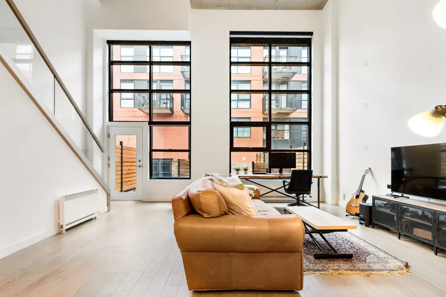 Bright living room with a brown leather sofa, wood floors, and tall windows overlooking a brick building. A desk, TV, and guitar are also visible.