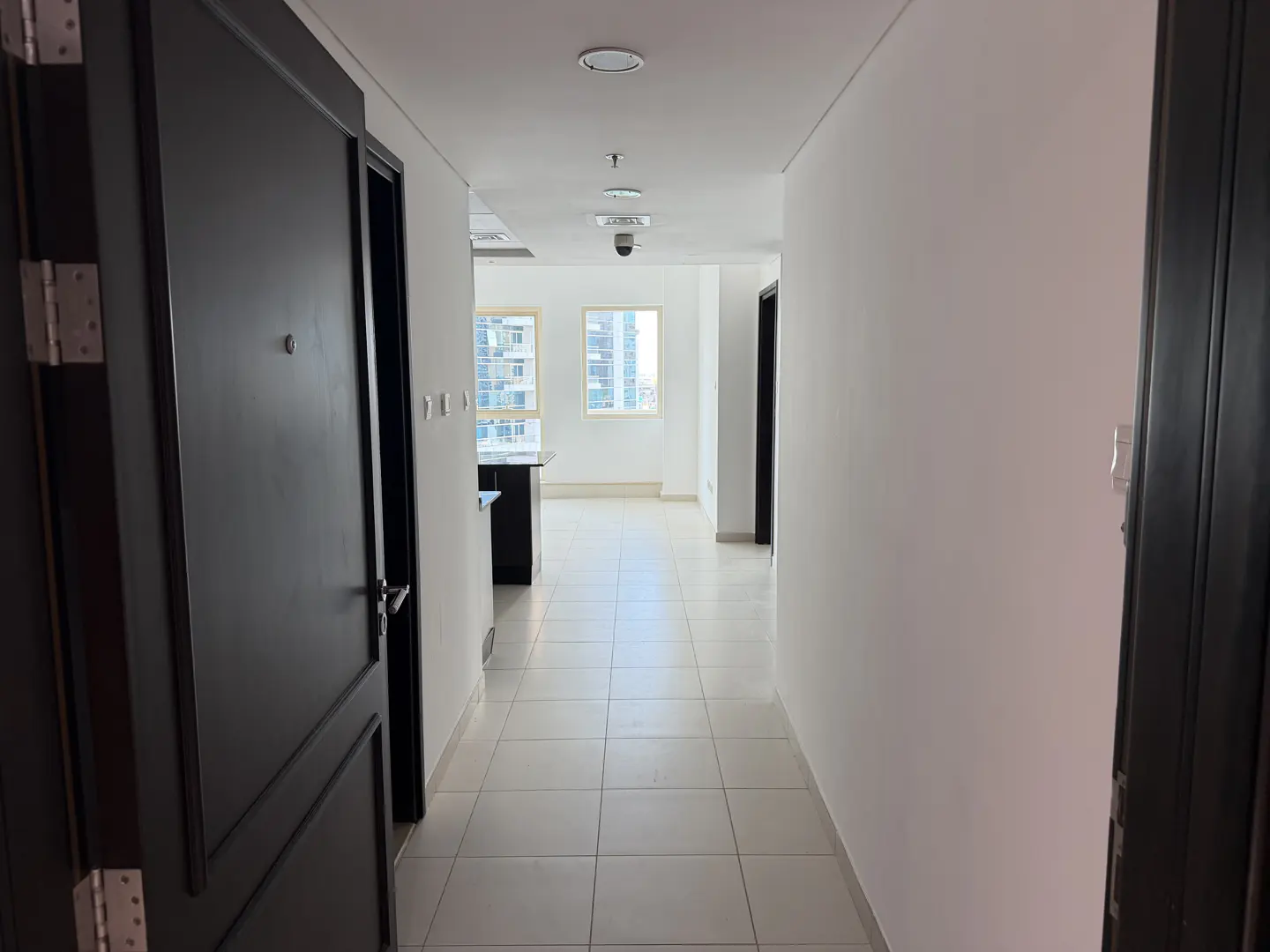 A bright hallway with white walls and tiled floors, viewed through an open dark door. A window at the end shows a city view.
