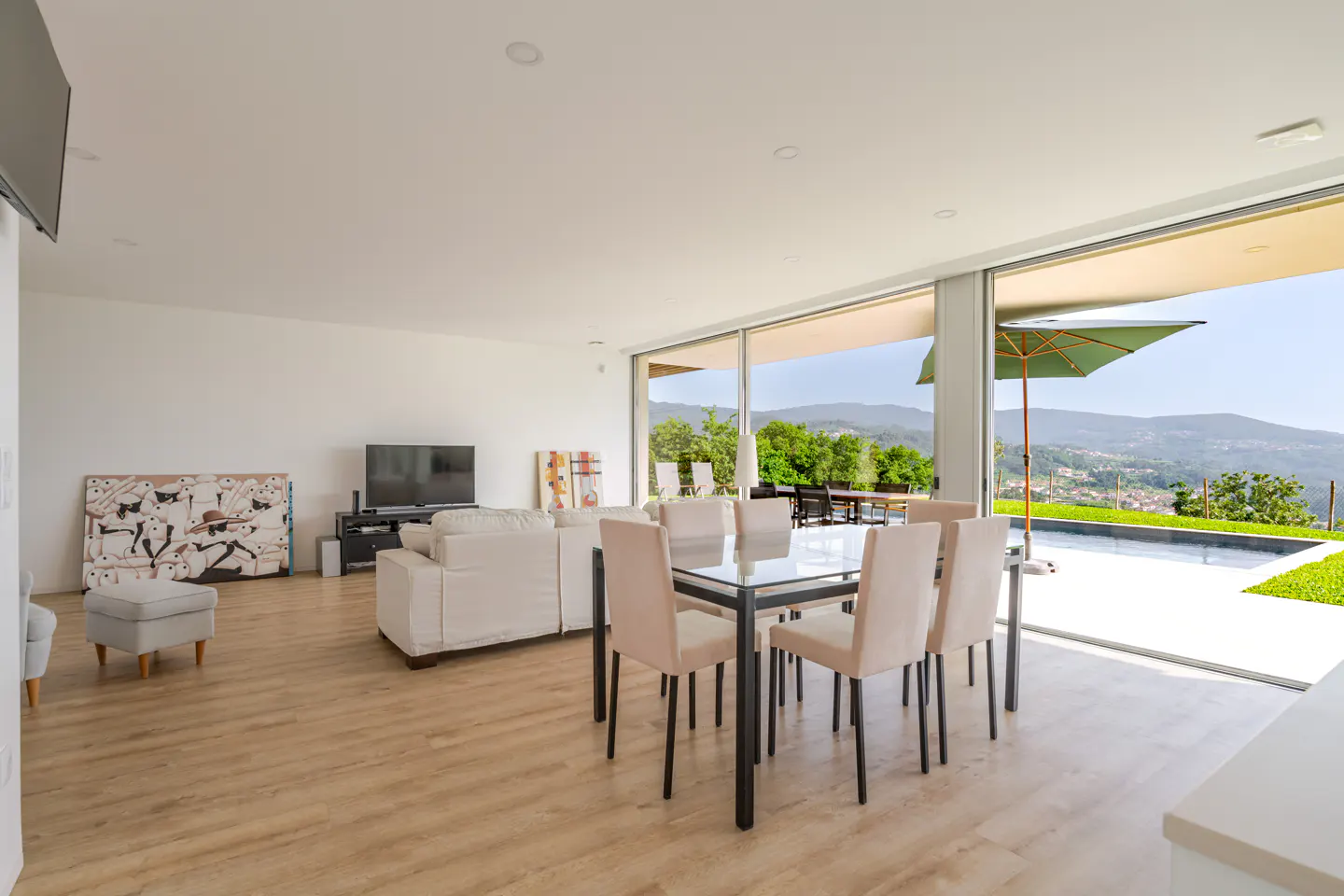 Modern open-plan living area with white sofa, glass dining table, and sliding doors opening to a pool and mountain view.