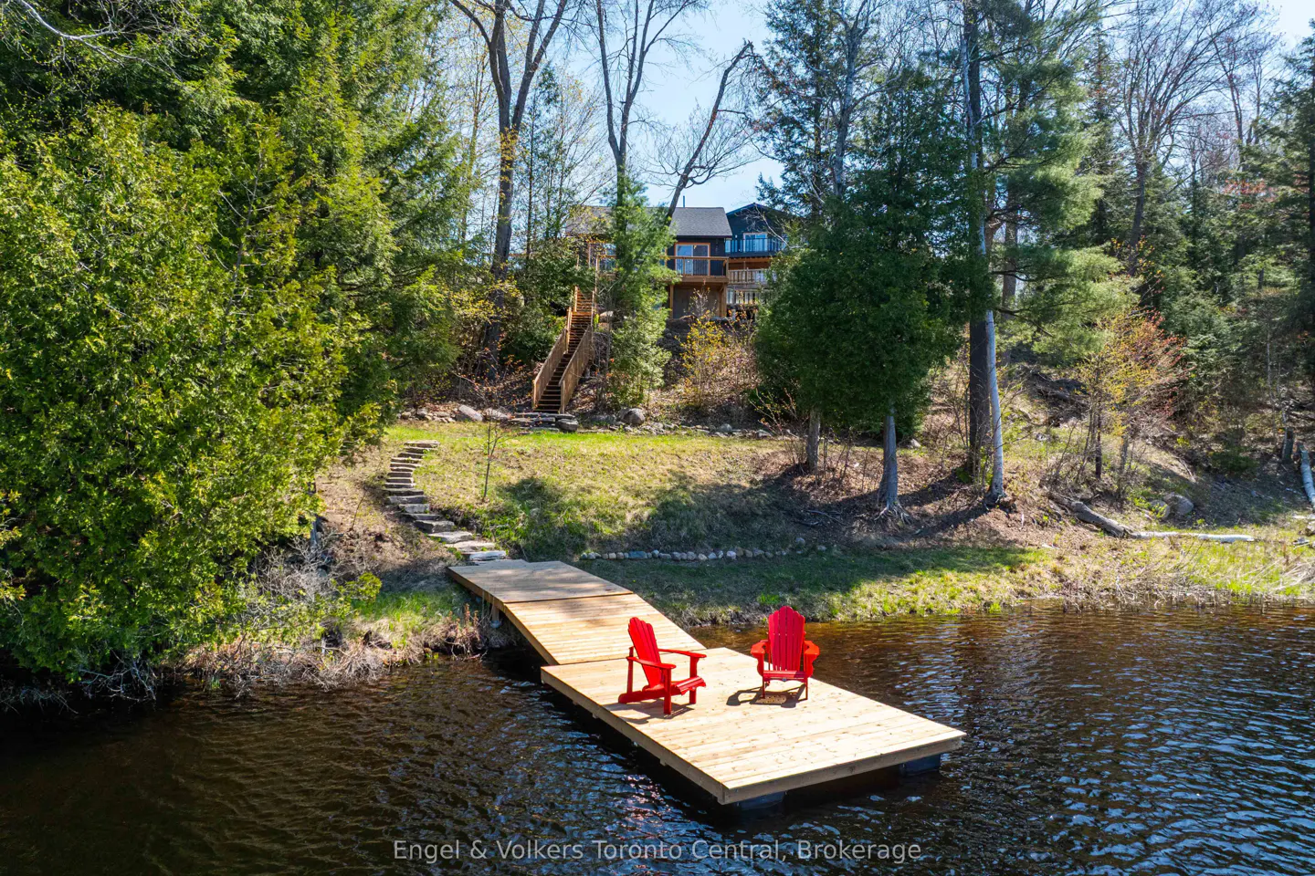 Waterfront property view: Two red Adirondack chairs sit on a wooden dock extending into a lake. Stone steps lead up to a house nestled among green trees.
