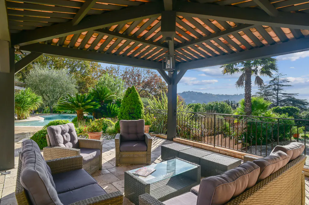 Covered patio with wicker furniture, gray cushions, and a glass table. Lush green landscape with a pool in the background.