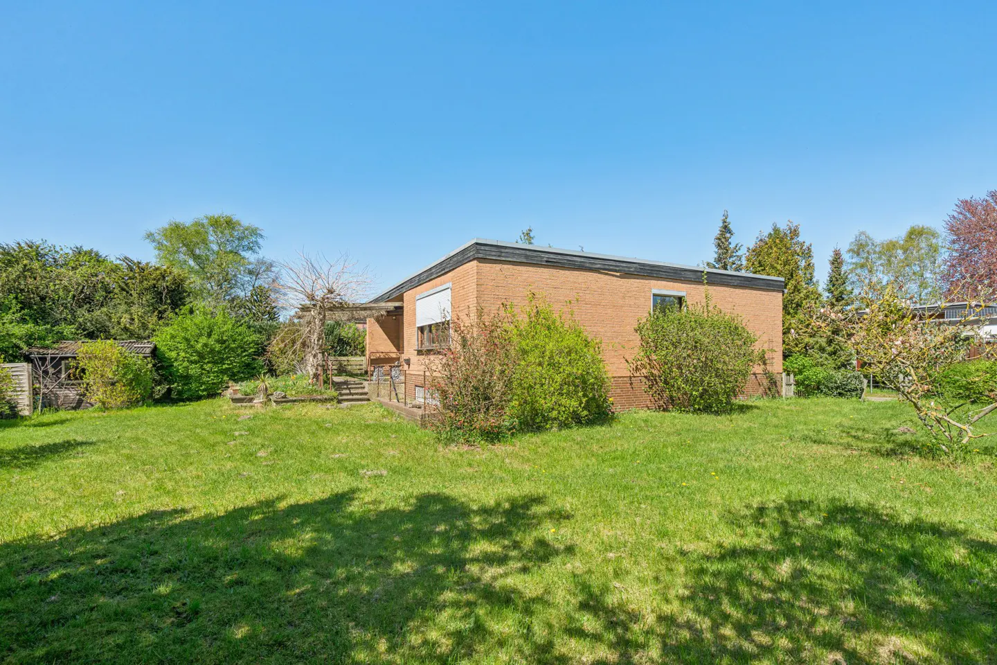 Backyard view of a single-story brick house with a green lawn and trees under a clear blue sky.