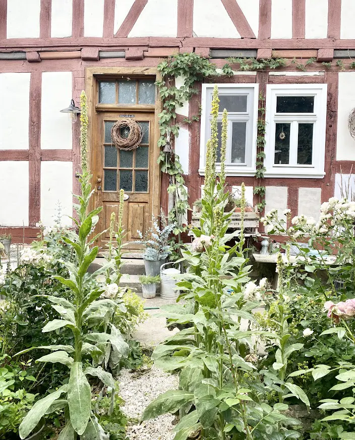 Charming half-timbered house with a wooden door, wreath, and lush garden. White walls, red beams, and green plants create a cozy, inviting scene.