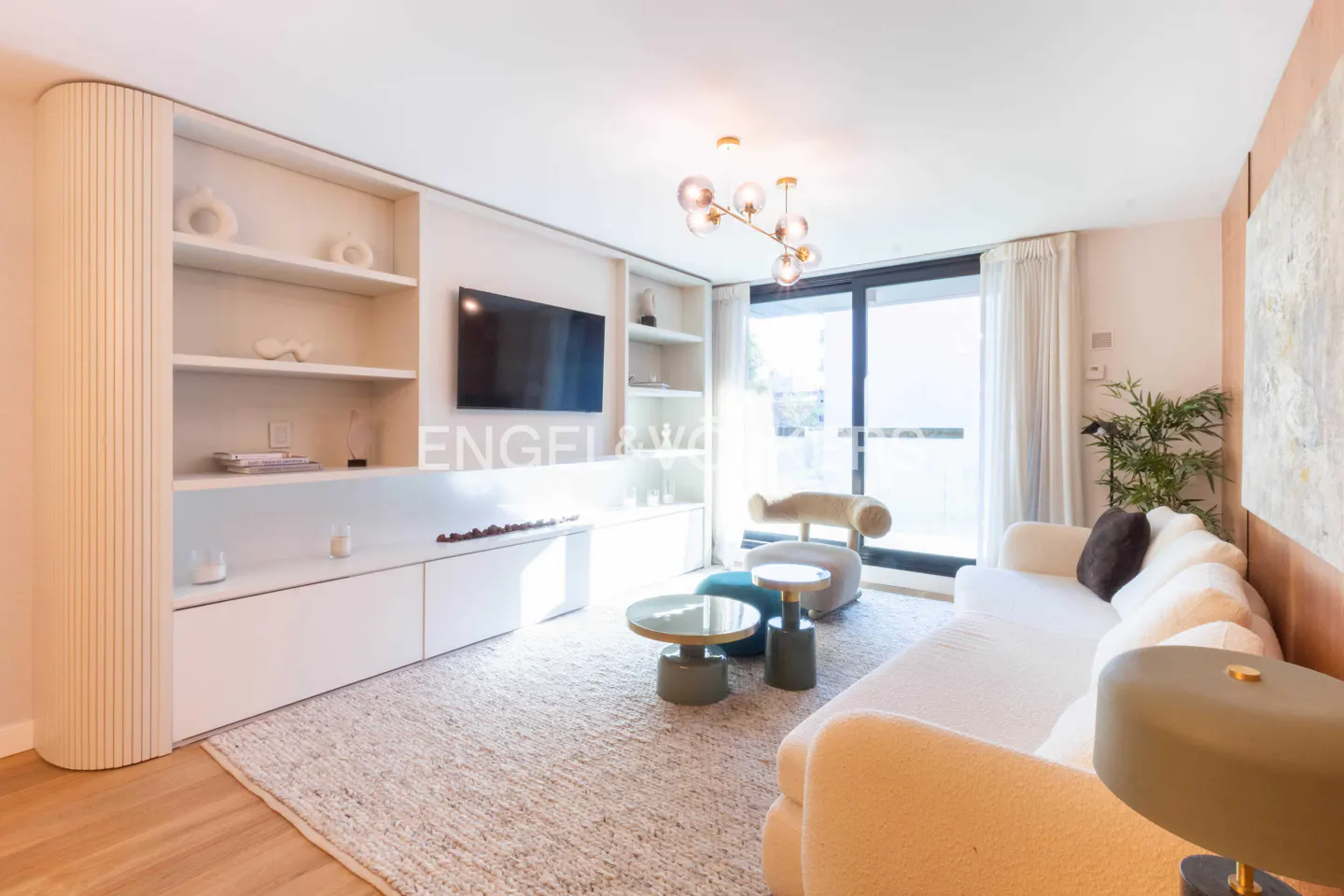 Bright living room with white sofa, shelves, and TV. A modern chandelier hangs above, and a large window lets in natural light.