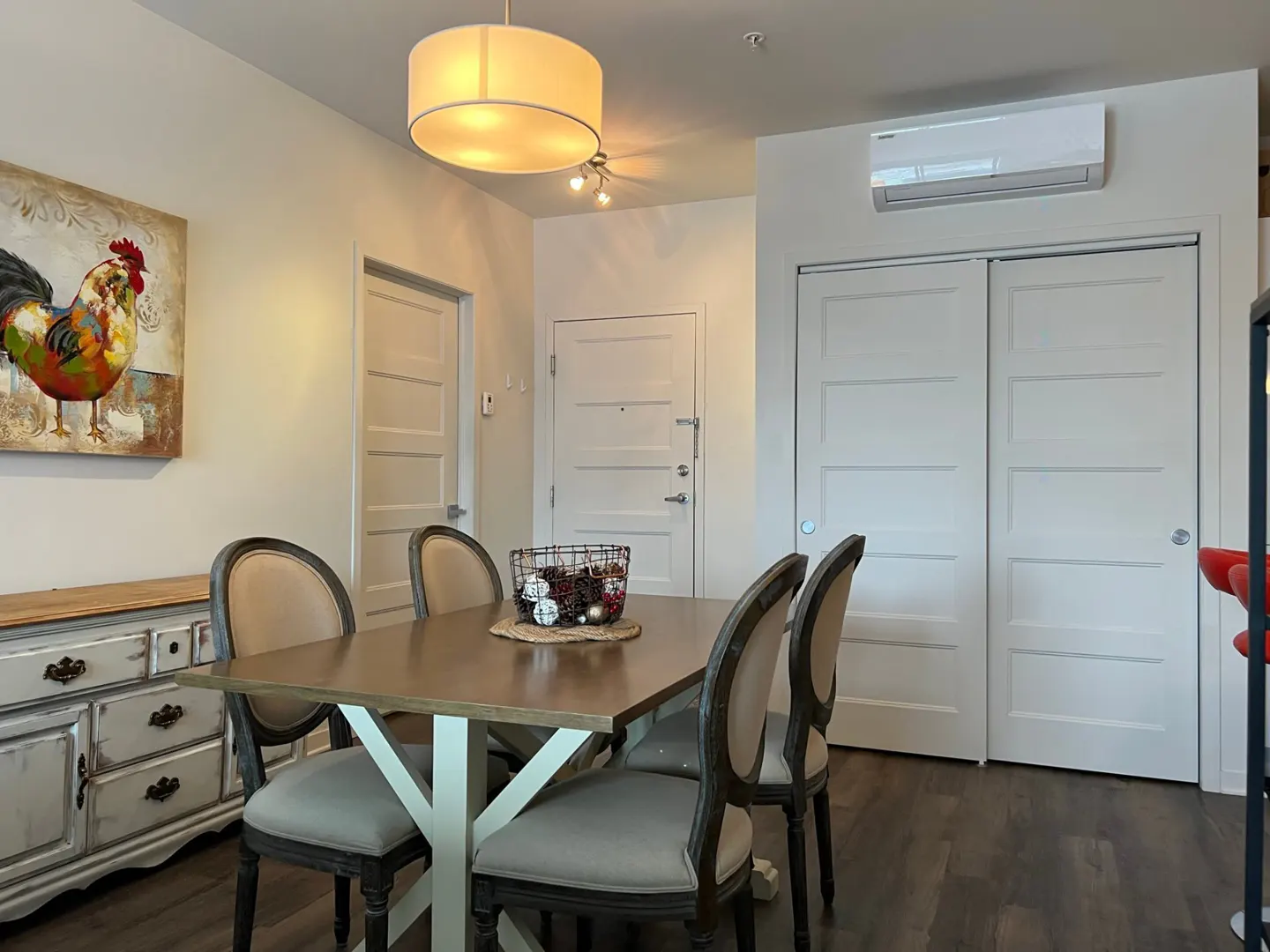 Dining area with a wooden table, six chairs, and a basket centerpiece. A colorful rooster painting hangs above a distressed sideboard. White walls and dark wood floors.
