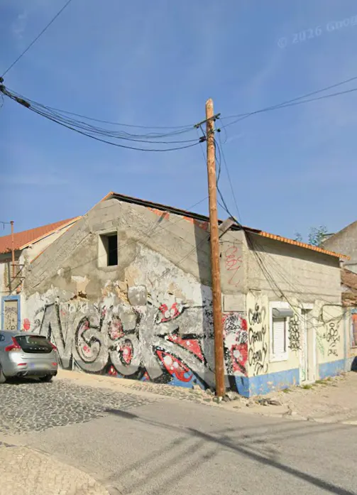 Exterior view of a rundown, graffiti-covered building with a silver car parked nearby on a cobblestone street.