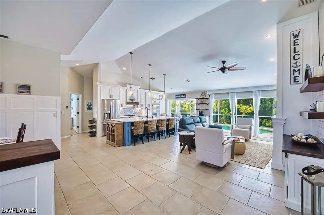 Open-concept living space with kitchen island, wicker stools, blue sofa, and white armchairs. Large windows overlook a pool. "Welcome" sign.