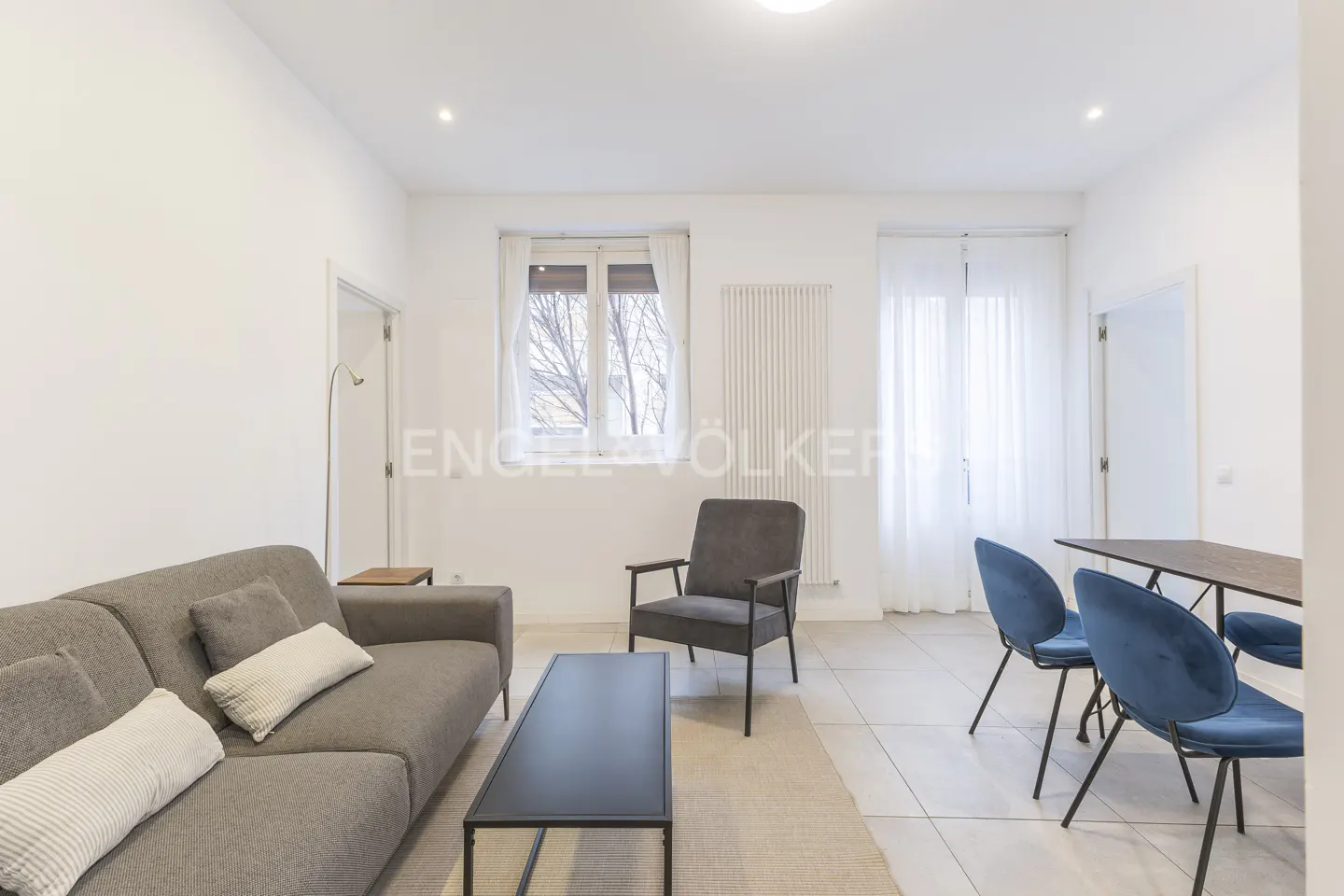 Bright living room with gray sofa, armchair, black coffee table, and dining table with blue chairs. White walls and tile floor.