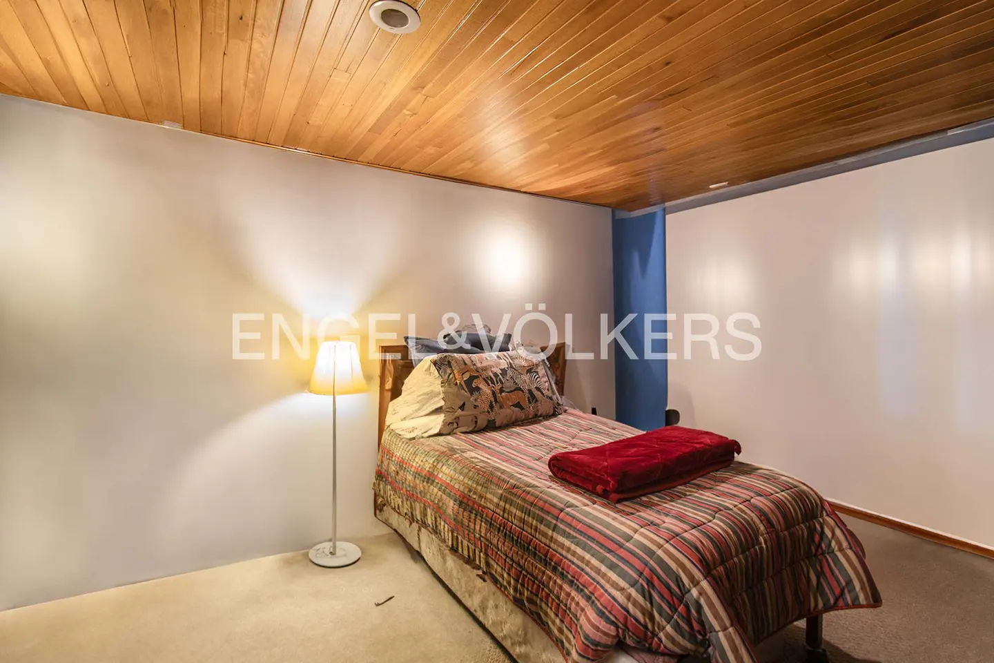 Bedroom with wood-paneled ceiling, white walls, and a bed with a red throw blanket. A floor lamp stands beside the bed.