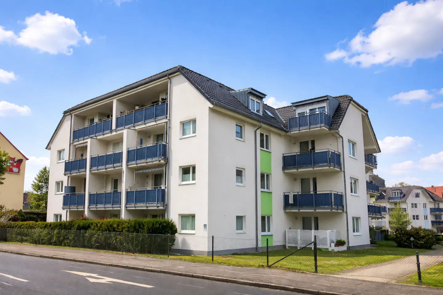 Three-story white apartment building with blue balconies under a blue, cloudy sky.