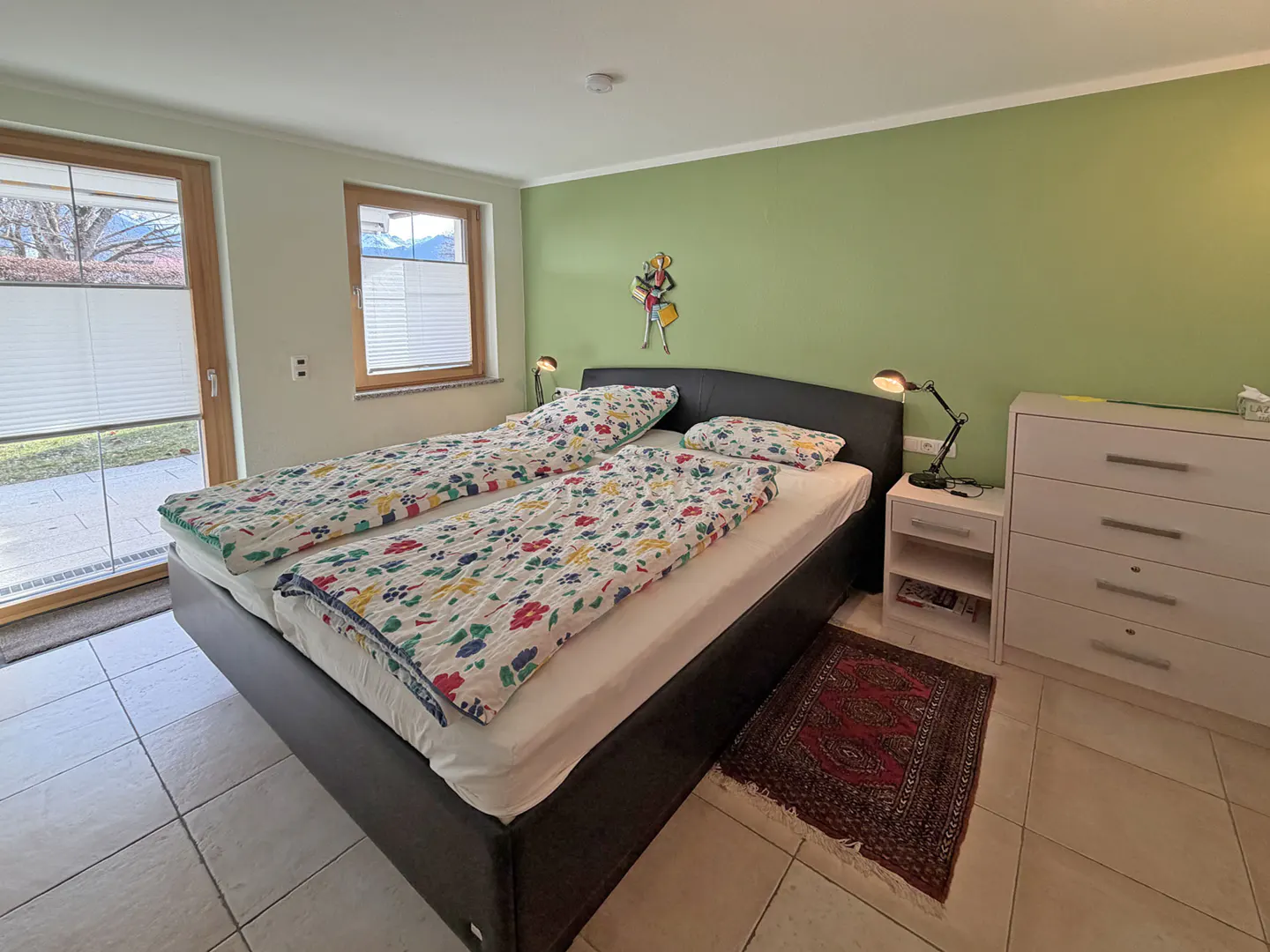 Bedroom with a queen bed, floral bedding, a green wall, and a white dresser. Windows with blinds are on the left.