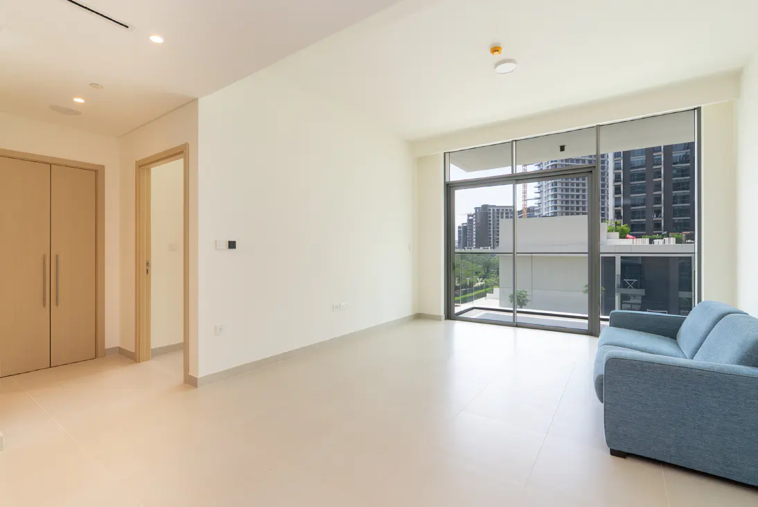 Bright, empty room with beige tile floor, white walls, and a blue sofa. Large windows overlook city buildings.