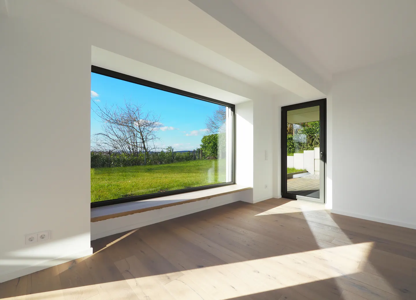 Bright, empty room with wood floors, white walls, and a large window showing a green field and blue sky. A black-framed door is slightly ajar.