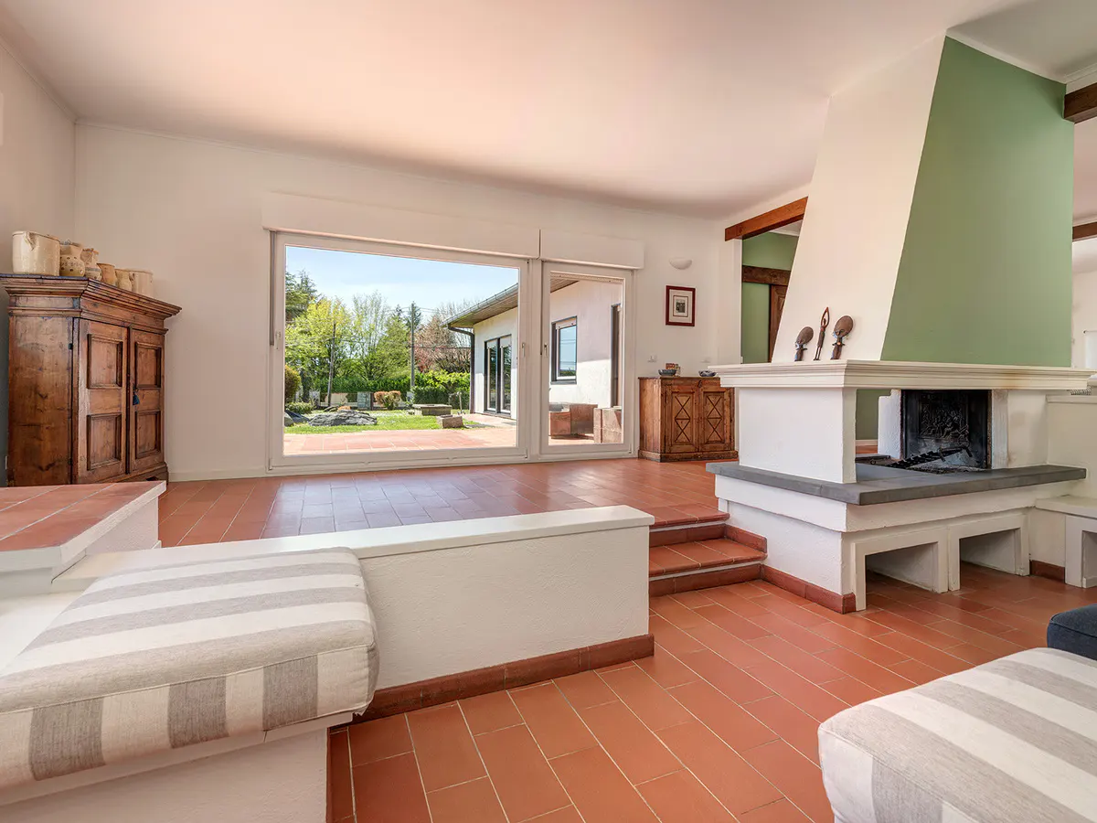 Bright living room with terracotta tile floor, white walls, and a large window overlooking a green lawn. A white fireplace with a green accent wall is visible.