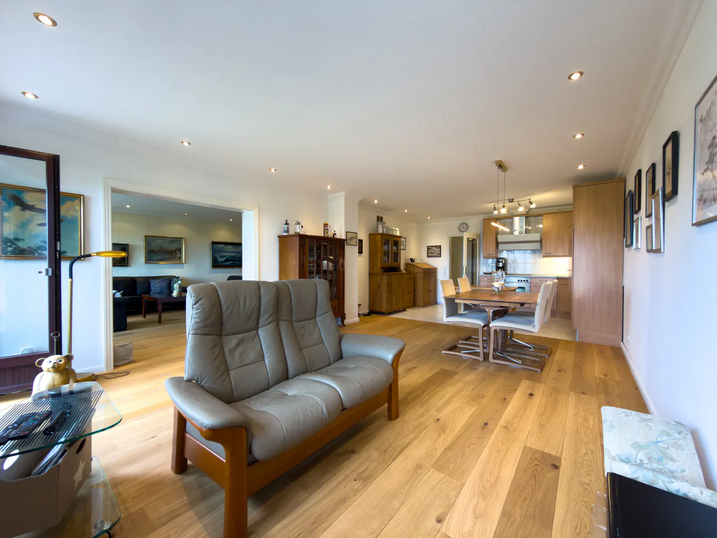 Open-concept living space with a gray leather sofa, wood floors, dining table, and kitchen in the background.
