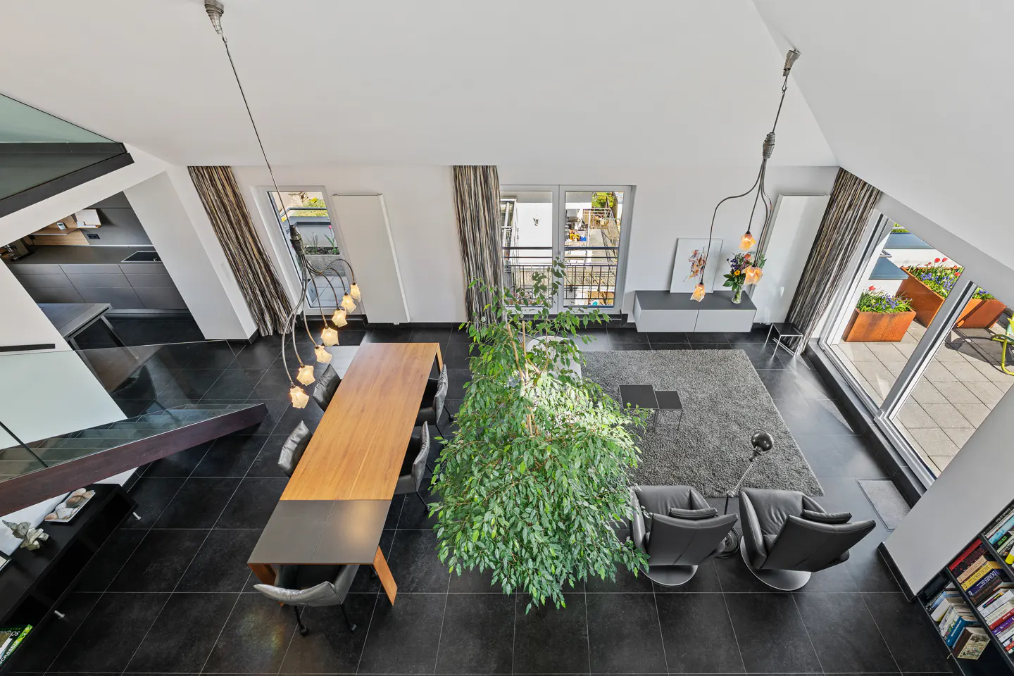 Overhead view of a modern living space with black tile floors, a wood table, and a large green plant. Balcony with flowers visible.