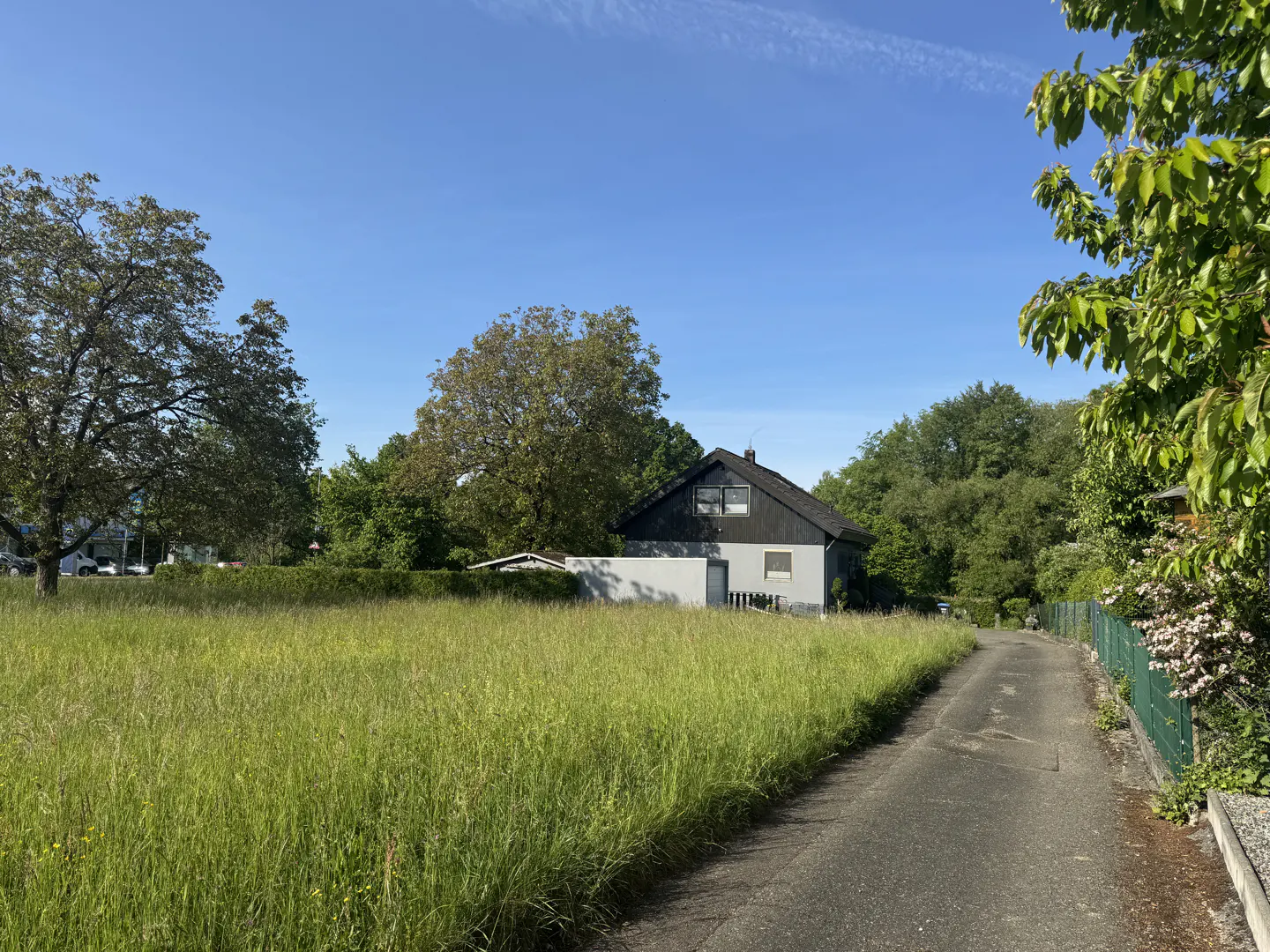A paved path leads past a large, grassy field toward a two-story house with a dark roof under a clear blue sky.