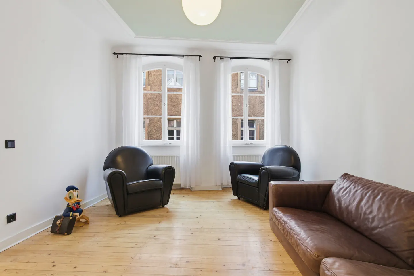 Bright living room with wood floors, white walls, and two black armchairs. A brown leather couch sits to the right, and a Donald Duck figure is on the left. Two windows with white curtains let in natural light.