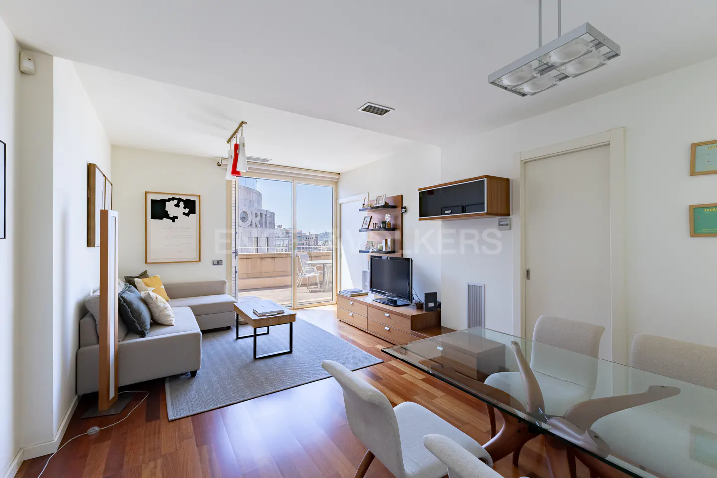 Bright living room with a gray sofa, wood floors, and a glass dining table. A balcony is visible through sliding glass doors.
