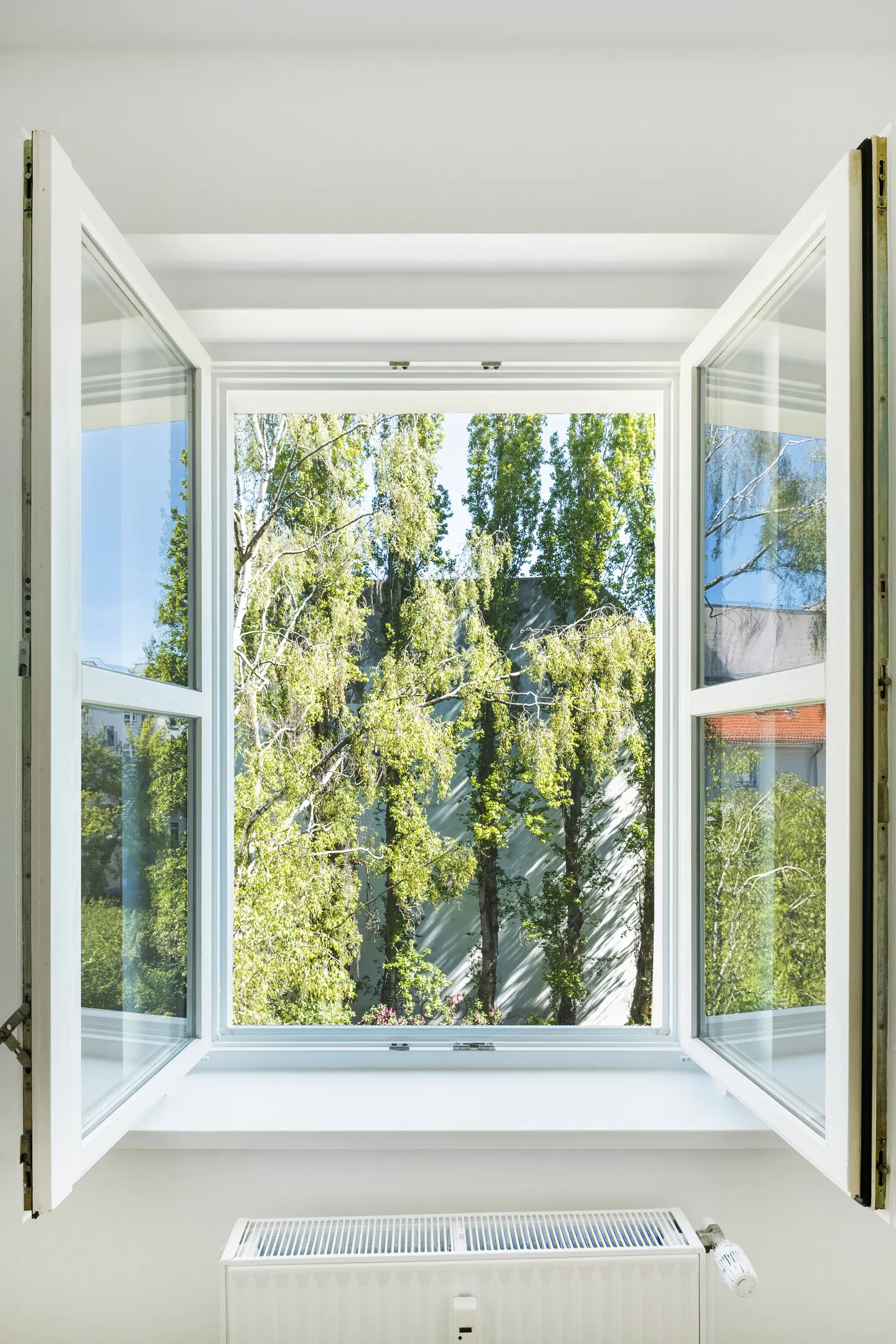 Open white window with trees and a building visible outside. A white radiator is below the window.