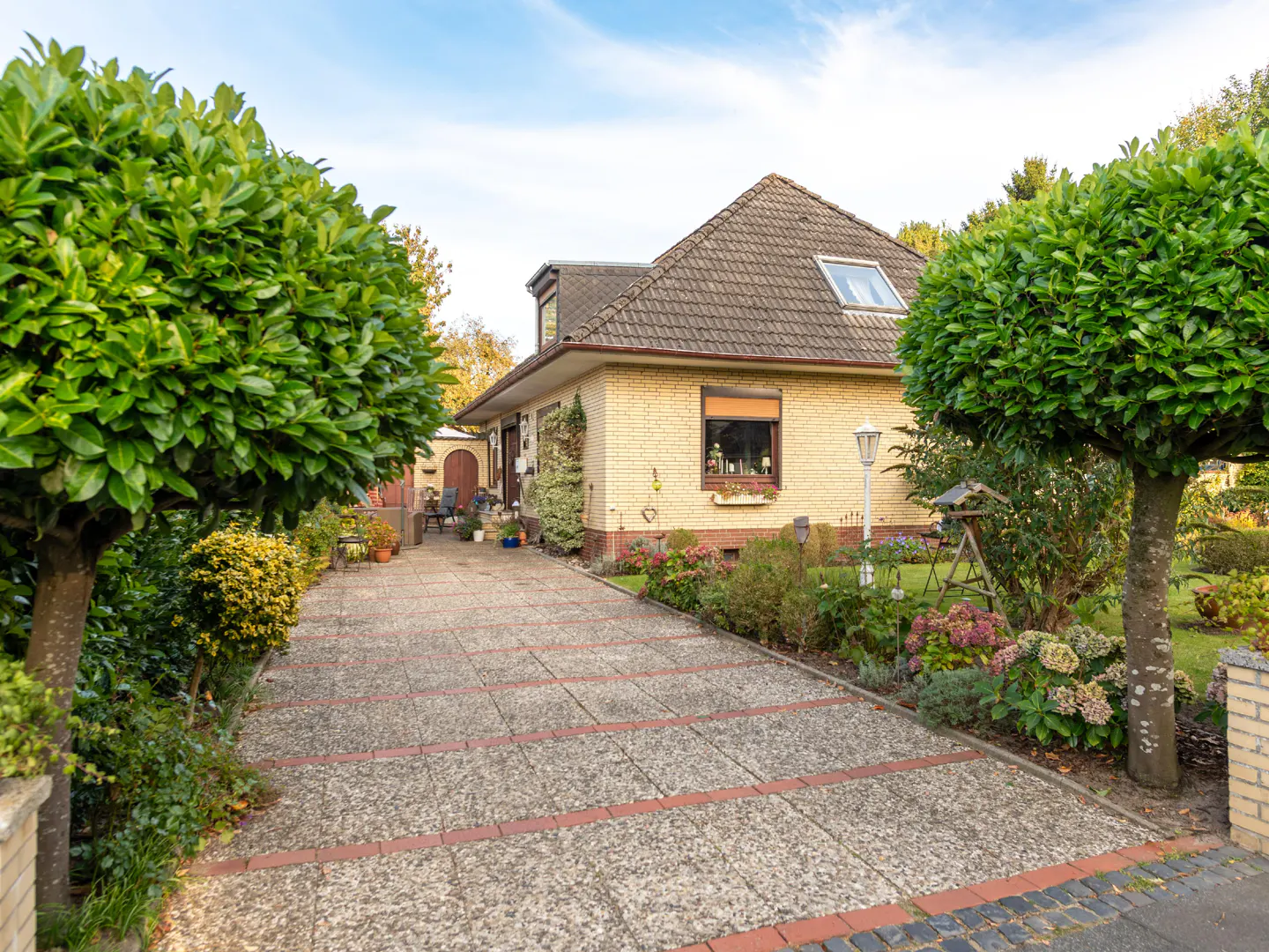 Exterior view of a one-story yellow brick house with a brown roof and a stone driveway lined with green trees and flowerbeds.
