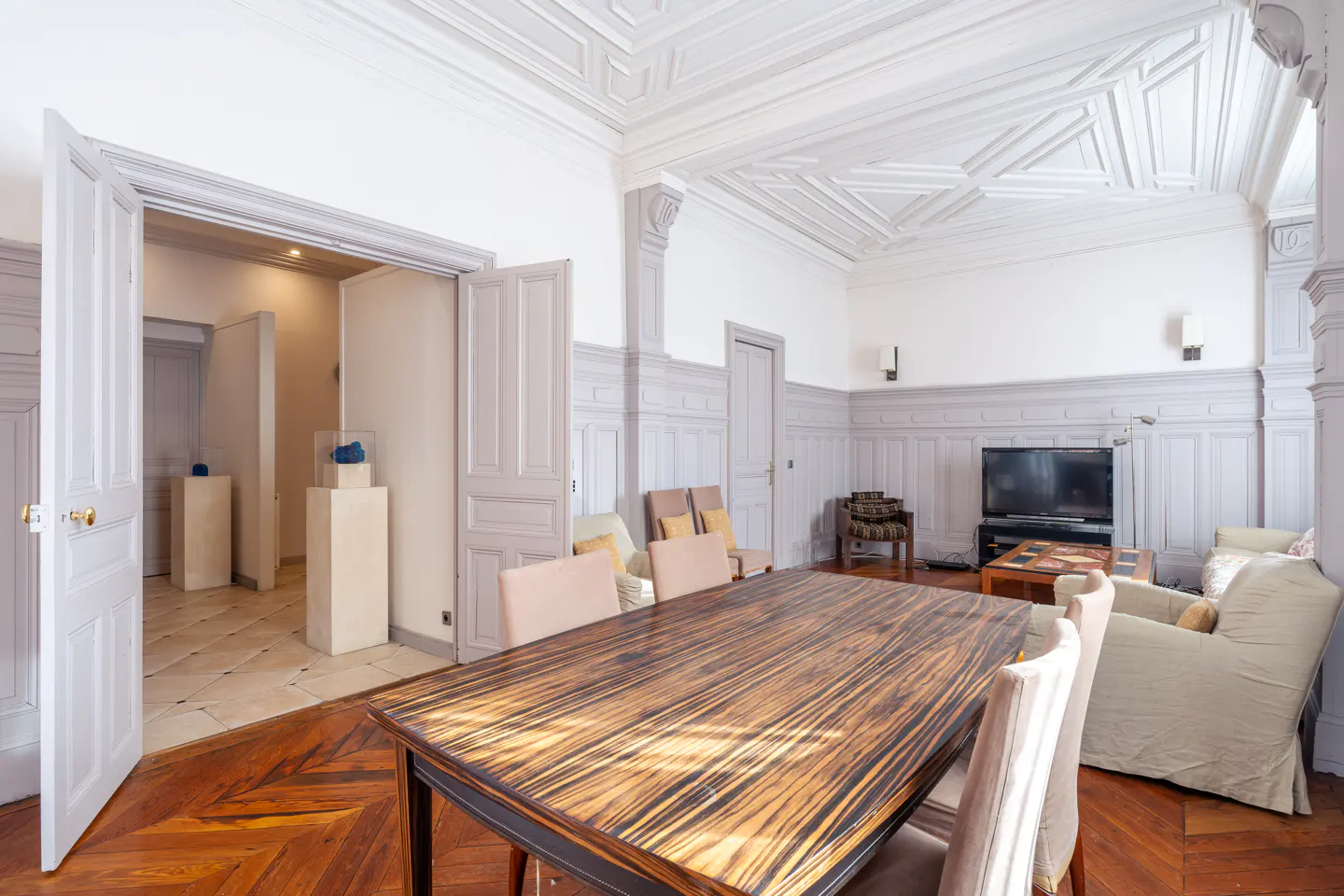 A bright dining room with a wood table, chairs, and herringbone floors. An open doorway leads to a hall with art displays.