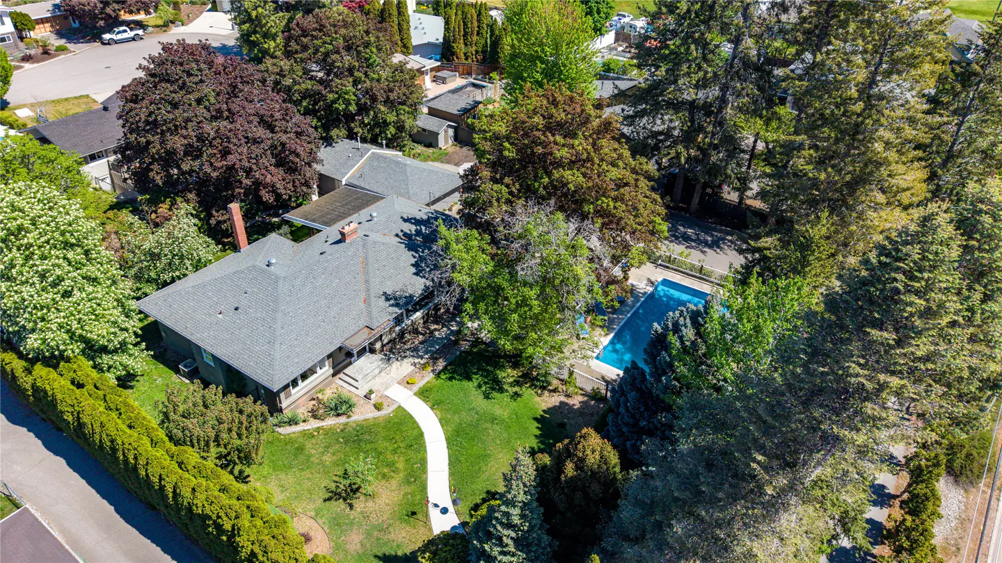 Aerial view of a house with a gray roof, green lawn, white walkway, and a blue swimming pool surrounded by trees.