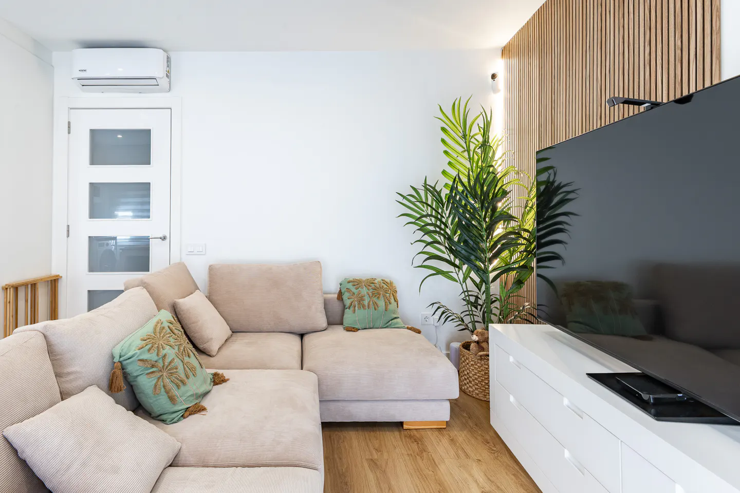 Bright living room with a beige sectional sofa, green pillows, a large TV, and a potted palm tree against a wood-paneled wall.