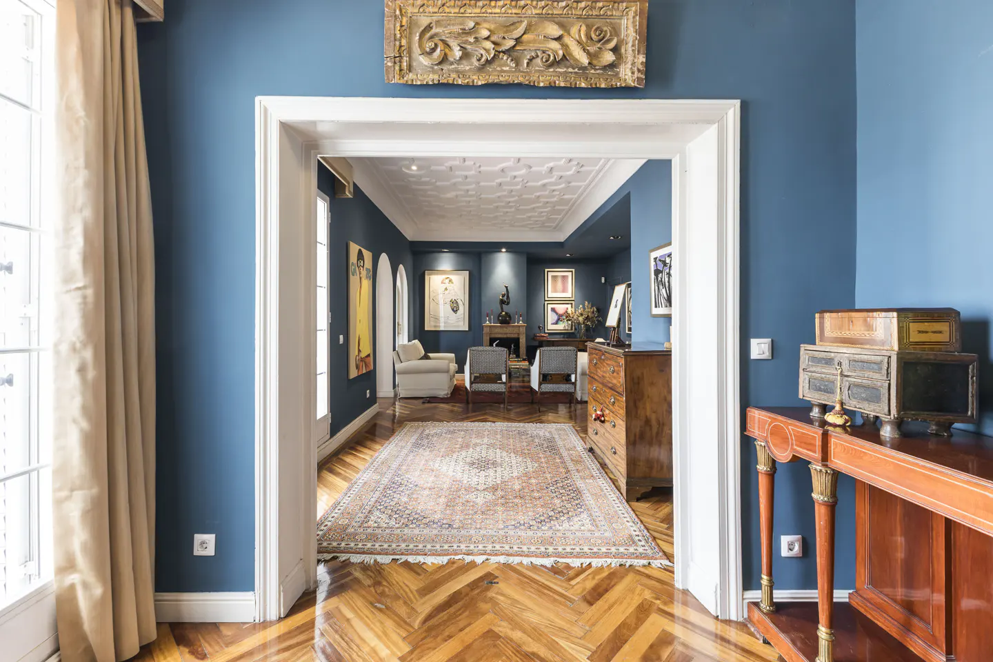 View through a doorway into a blue room with a patterned rug, wood floors, and furniture. A gold ornate frame hangs above the doorway.