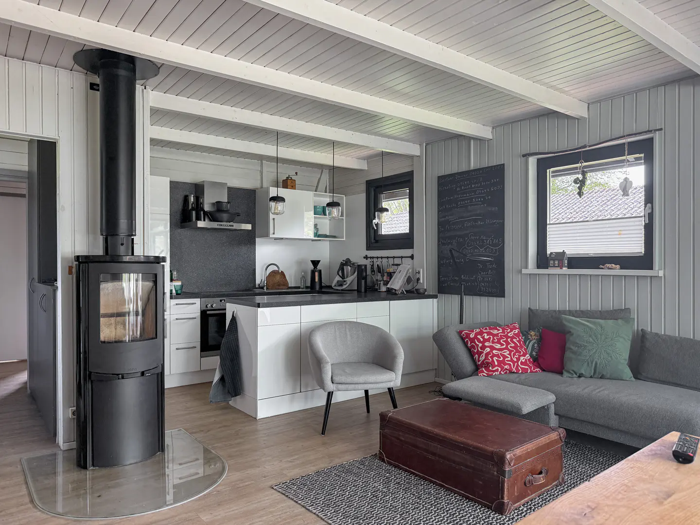 Modern open-plan living room with a black wood stove, white kitchen, grey sofa, and a vintage leather trunk coffee table.