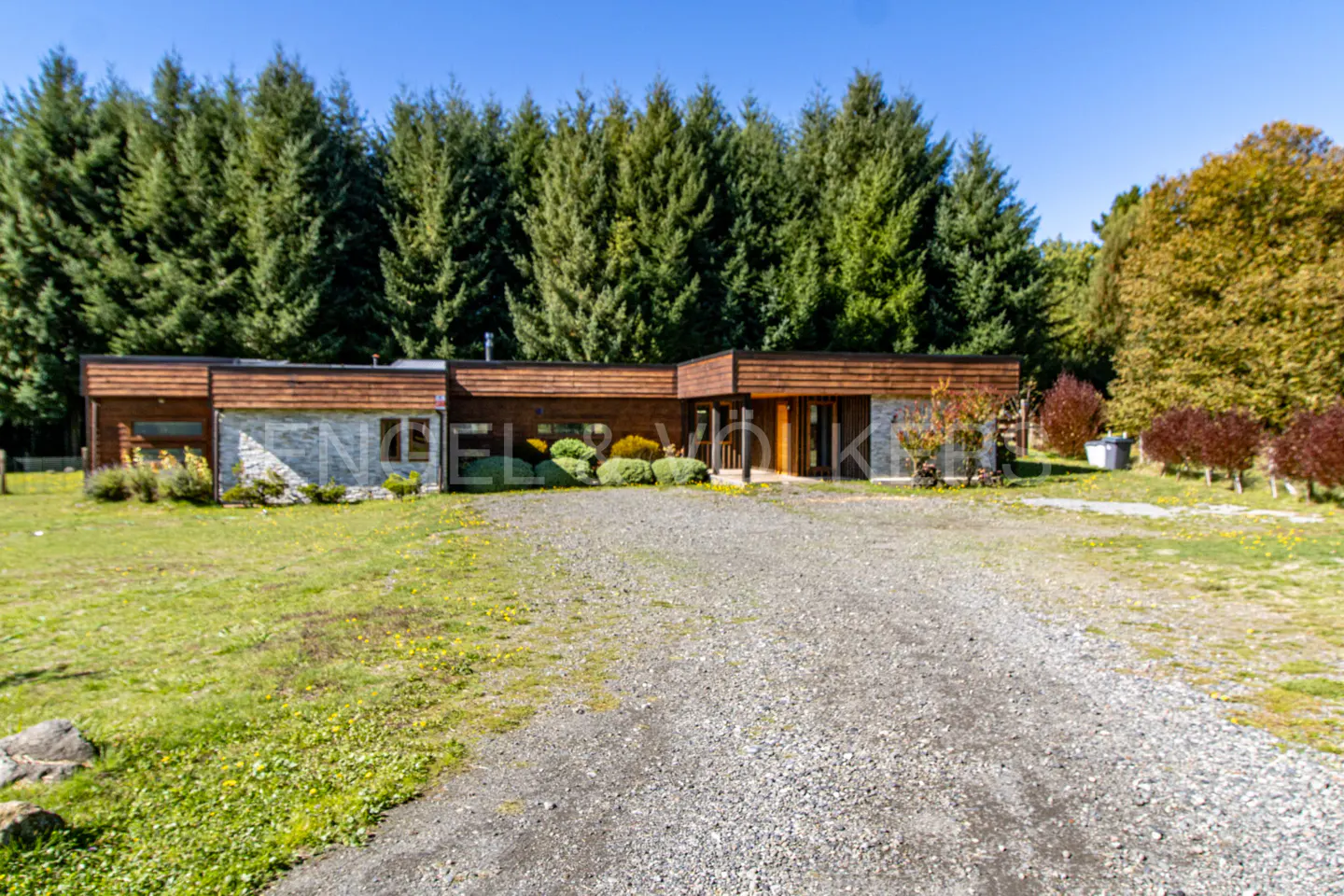 A single-story house with a wood and stone facade, surrounded by green grass and tall trees under a blue sky.