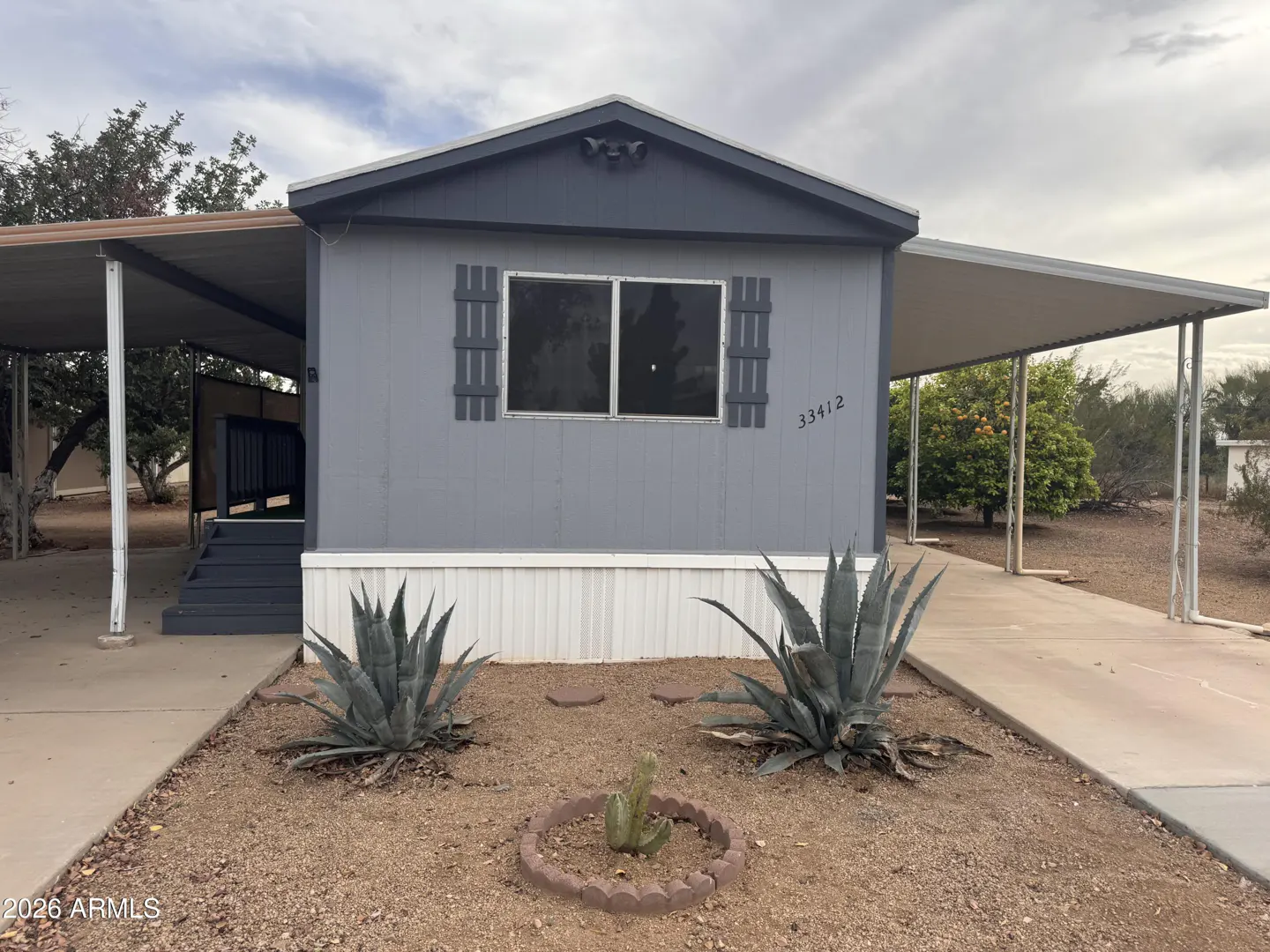 A gray single-story home with dark gray trim and shutters, with desert landscaping and carports on both sides.