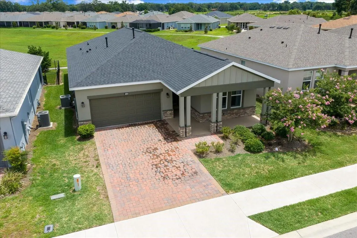 Aerial view of a one-story house with a gray roof, a brick driveway, and a covered porch with columns.