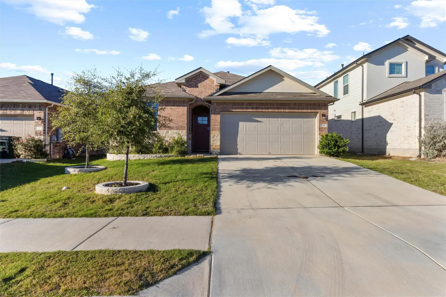 A single-story brick house with a beige garage door and a brown front door. A concrete driveway leads to the street. Green lawn and trees in the front yard.