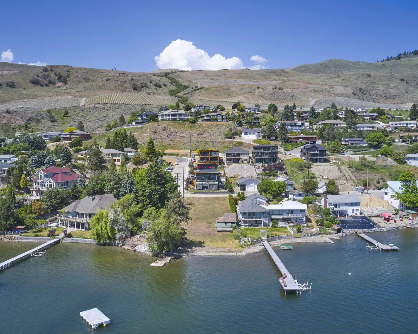 Waterfront homes with docks line a lake, backed by a hillside neighborhood under a blue sky with a puffy white cloud.
