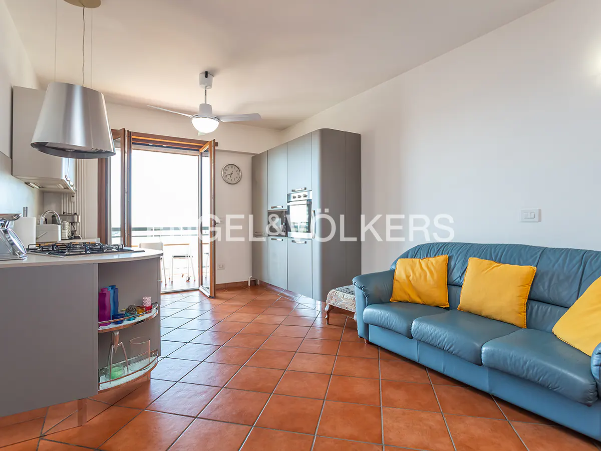 Open-concept living space with blue sofa, yellow pillows, terracotta tile floor, and kitchen with gray cabinets. Balcony in background.