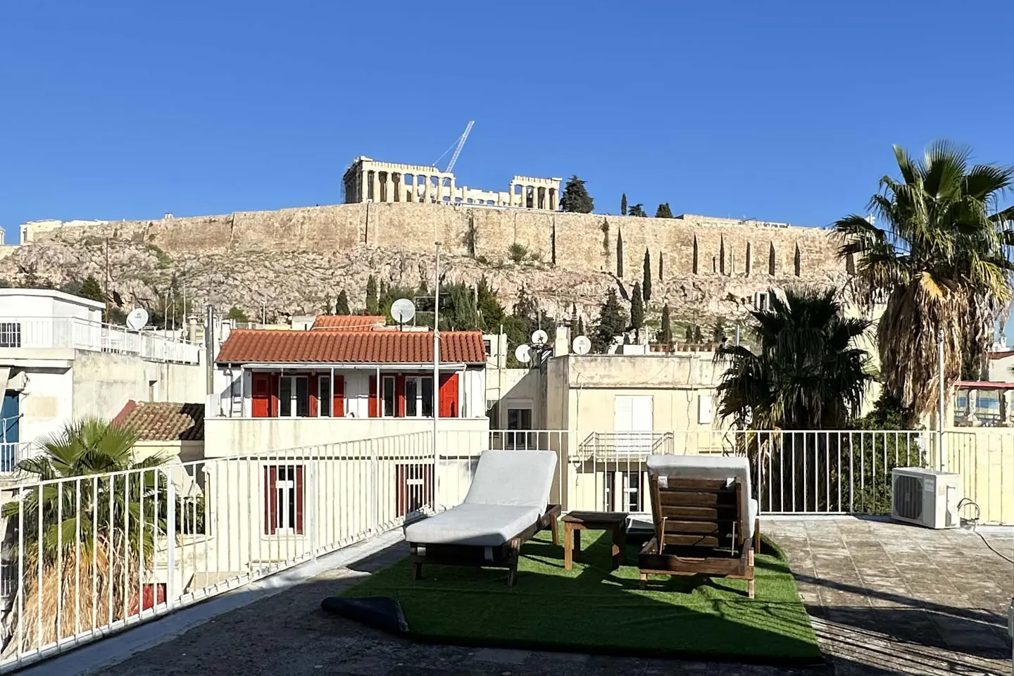 Rooftop view of Athens, Greece, with lounge chairs on artificial grass. The Acropolis sits atop a hill in the background.