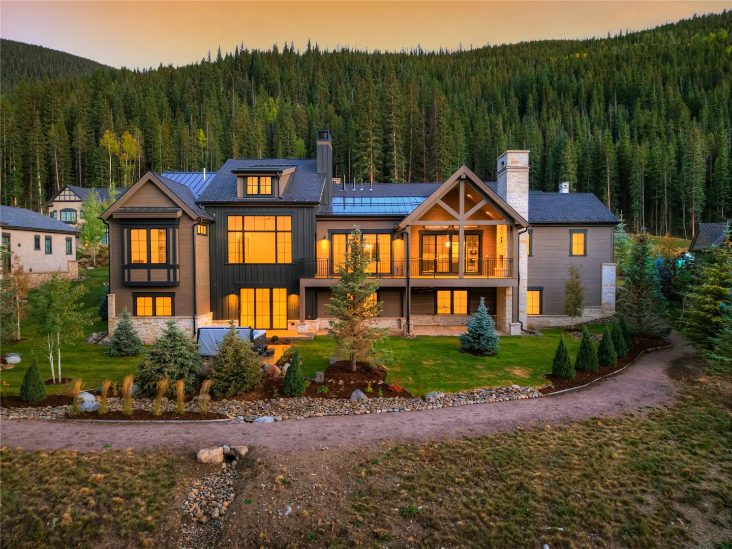 Luxury mountain home with large windows, brown siding, and a stone chimney, set against a backdrop of dense evergreen forest.