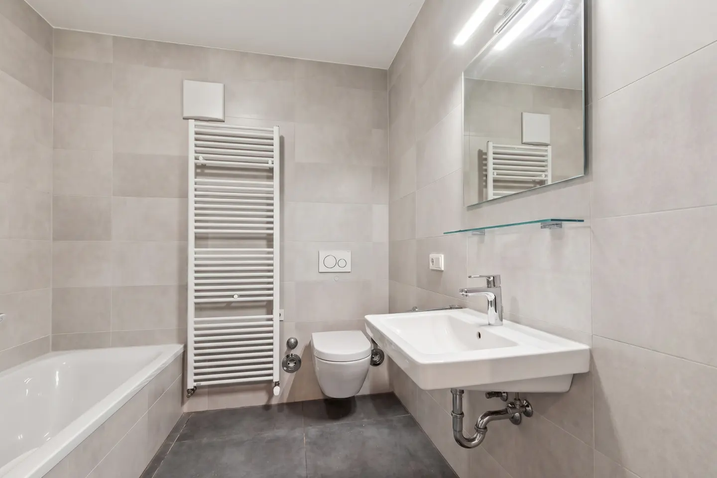 Modern bathroom with beige tiles, dark gray floor, white sink, toilet, bathtub, and towel warmer. A mirror hangs above the sink.