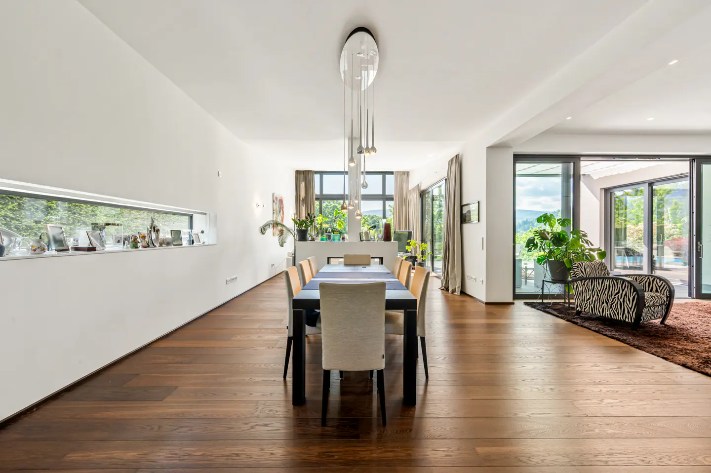Modern dining room with dark wood floors, a long table, pendant lighting, and a zebra-print chair in the living area.