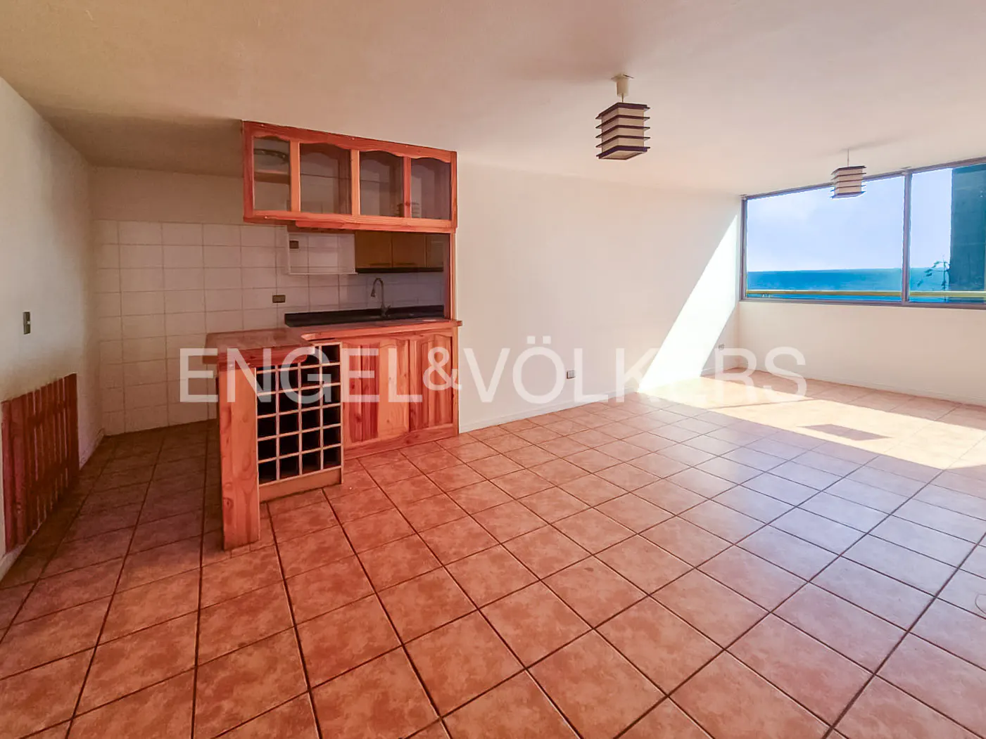 A bright, empty room with tile floors, a wooden kitchen counter, and a view of the ocean.