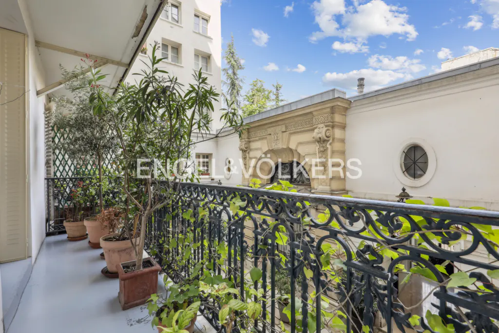 A Parisian balcony with potted plants and a black wrought iron railing. A building with a round window is in the background.