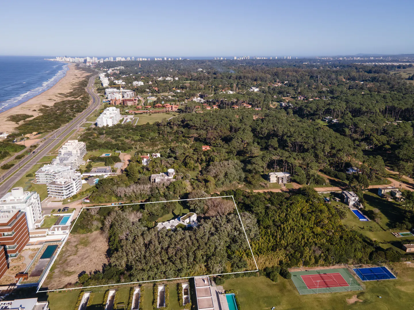 Aerial view of a property outlined in white, near a beach, trees, and tennis courts.