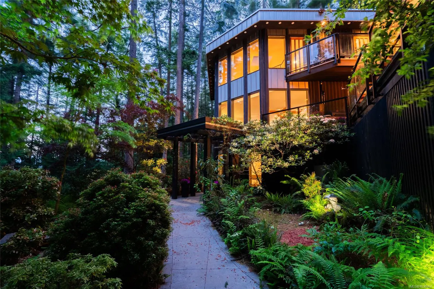 Exterior view of a modern house with large windows, a balcony, and a stone path surrounded by lush greenery and tall trees.