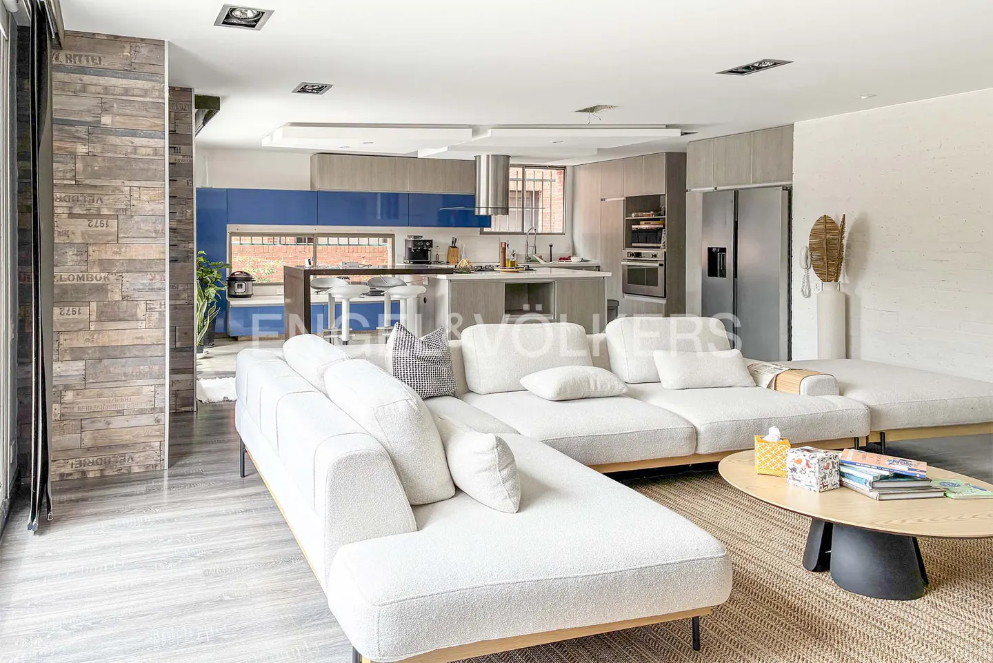 Open-concept living space with a white sectional sofa, wood coffee table, and a kitchen with blue cabinets in the background.